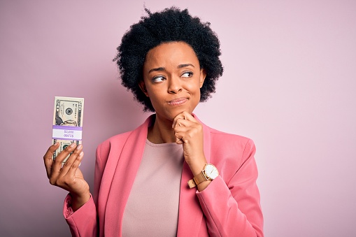 Young African American afro businesswoman with curly hair holding dollars banknotes serious face thinking about question, very confused idea